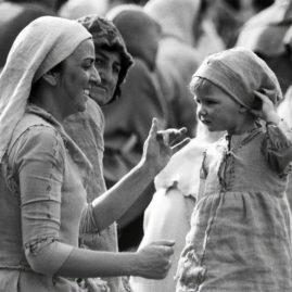 A smiling woman dressed as a medieval peasant costume is playing with her small child to distract her from the noise. Behind them an older woman is looking at them, or at the camera, with a slight frown.