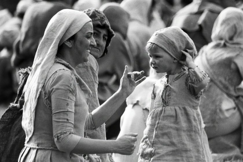 A smiling woman dressed as a medieval peasant costume is playing with her small child to distract her from the noise. Behind them an older woman is looking at them, or at the camera, with a slight frown.