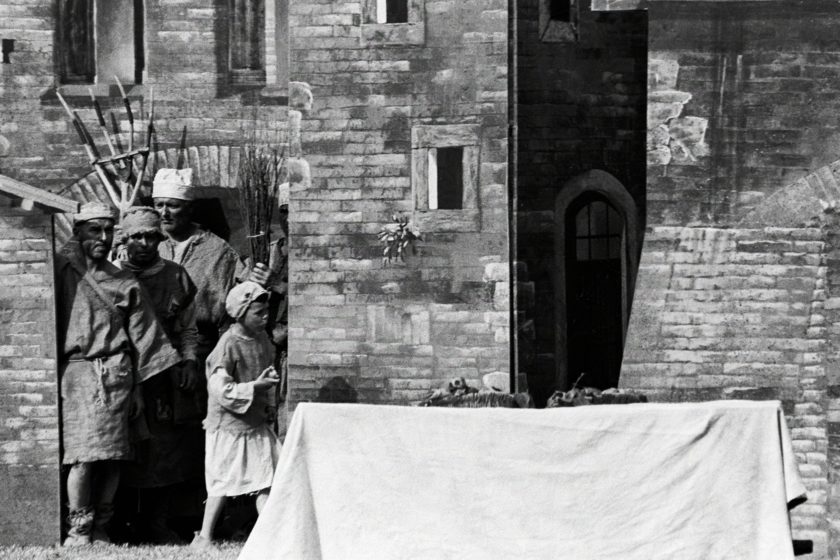 Men dressed in medieval garb are huddling against the scenery of amedieval town. A single kid is reaching otu towards the table in front, probably trying to get some food.