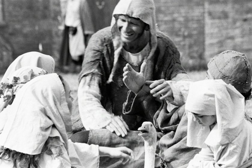 A man in medieval garb is playing with a few kids. The head of an alarmed-looking goose sticks out in front of the picture.