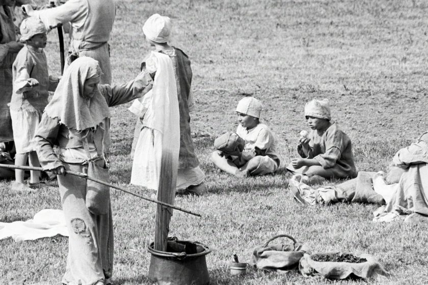 A woman in medieval garb is dyeing cloth in a pot. Behind her are a few kids, one of whom is eating an apple.