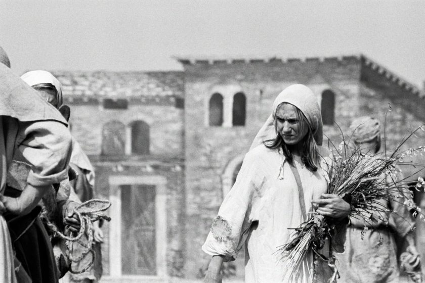 A woman in medieval garb is carrying a bunch of flowers. she is looking to the lower right, seemingly annoyed at what she's seeing.