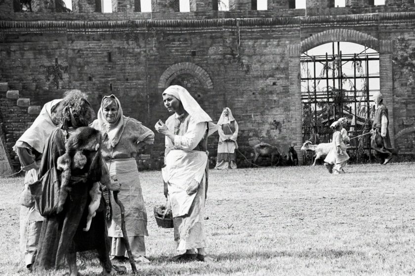 A group of women in medieval garb are gossiping with each other. In the background are two goard herders, by a piece of scenery that looks like a wall with an iron gate.